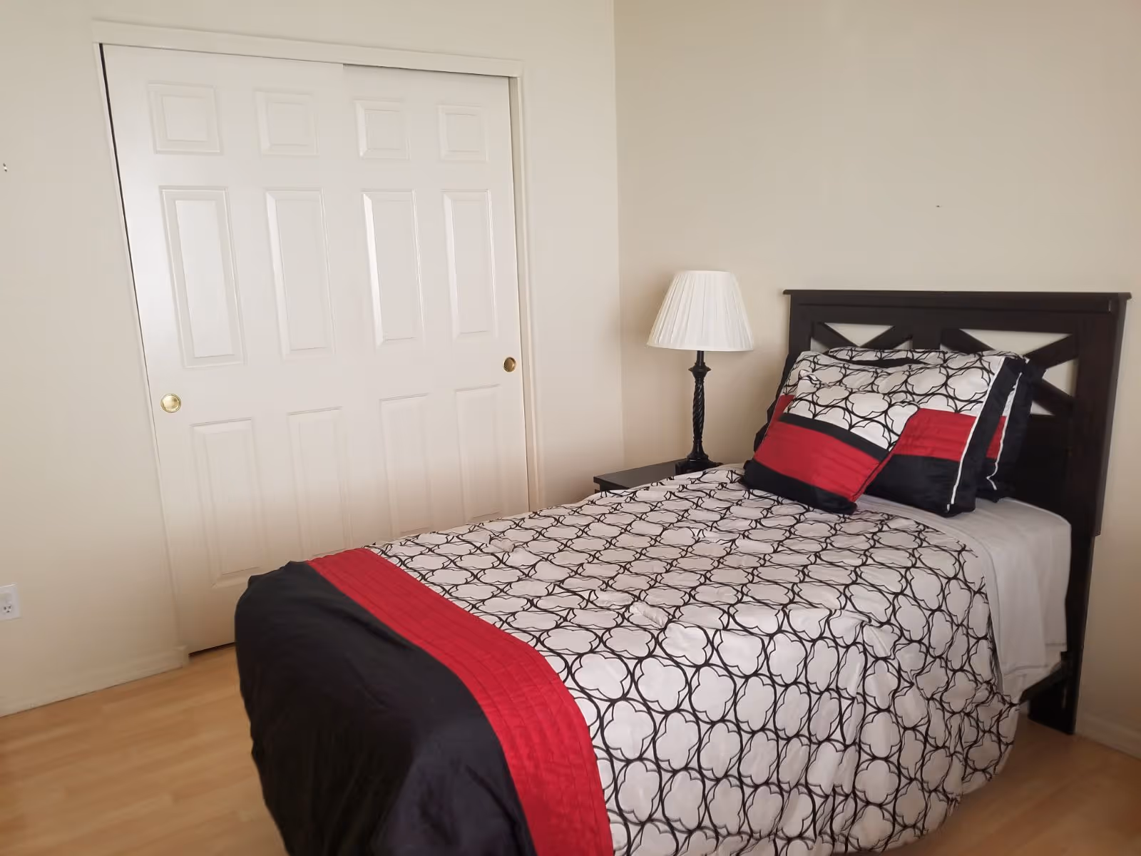 A neatly made single bed with a black, white, and red patterned comforter and matching pillows in a simple bedroom. Next to the bed is a small black nightstand with a white lampshade. Behind the bed is a plain beige wall, and to the left is a closed white double-door closet.
