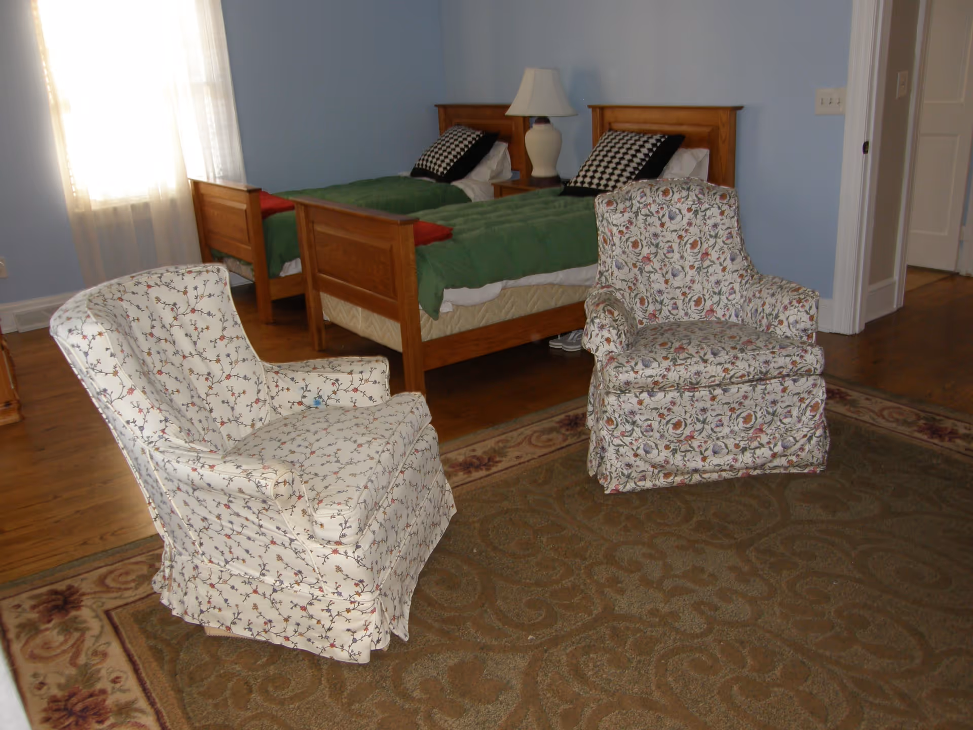 A bedroom with two twin beds featuring wooden frames and green bedding. Between the beds is a nightstand with a white lamp. In the foreground, there are two upholstered armchairs with floral patterns on a large area rug. The room has light blue walls and a window with sheer white curtains letting in natural light.