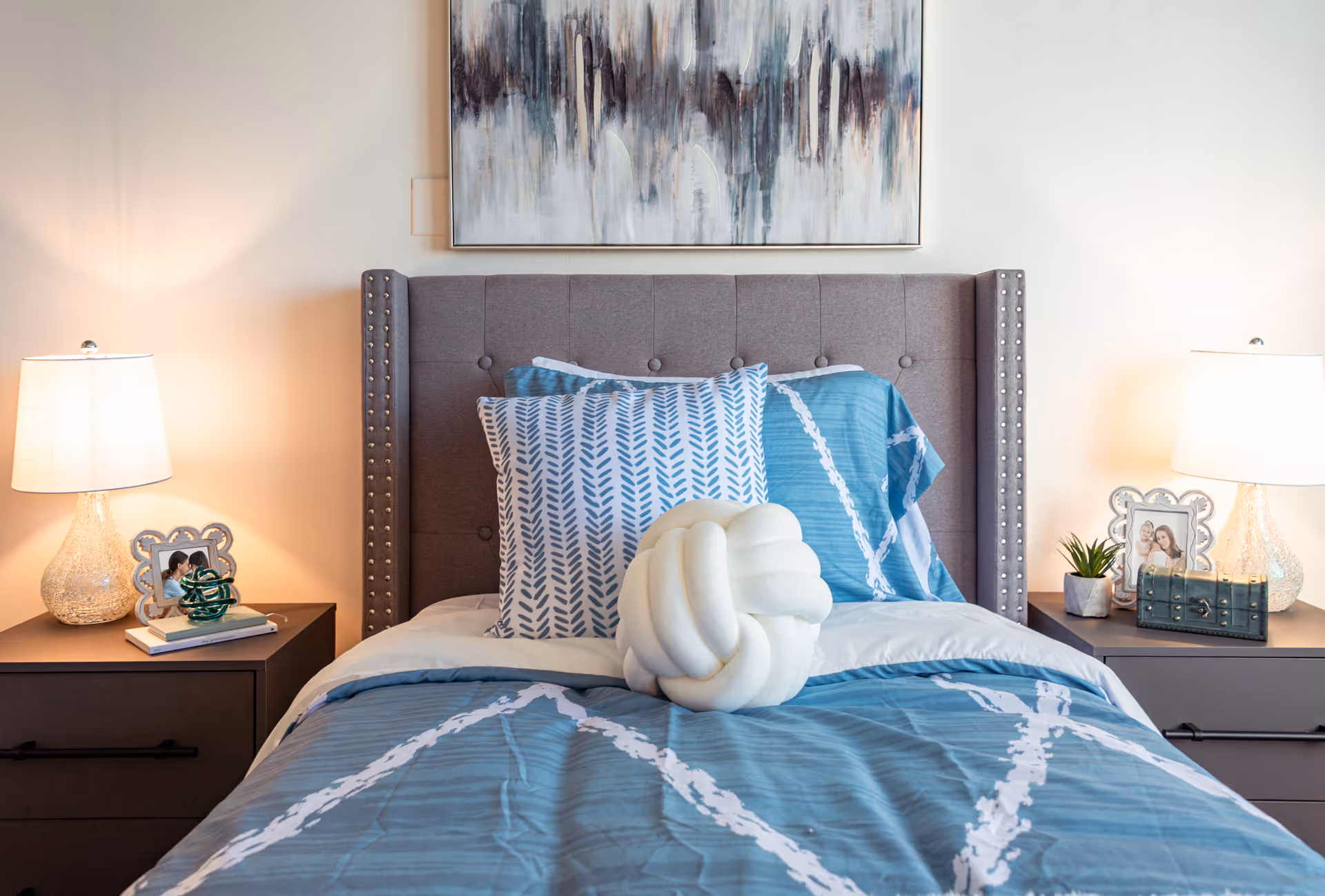 A neatly made bed with a gray upholstered headboard, blue and white patterned bedding, and decorative pillows. There are two bedside tables, each with a lamp, framed photos, and small decorative items. A large abstract painting hangs on the wall above the bed.