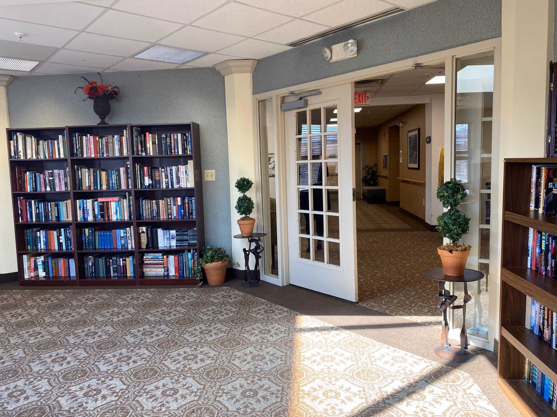 A quiet reading or lounge area in a senior living facility with two dark wood bookshelves filled with books, a patterned carpet, and two potted topiary plants on small round tables near a glass-paned door leading to a hallway.