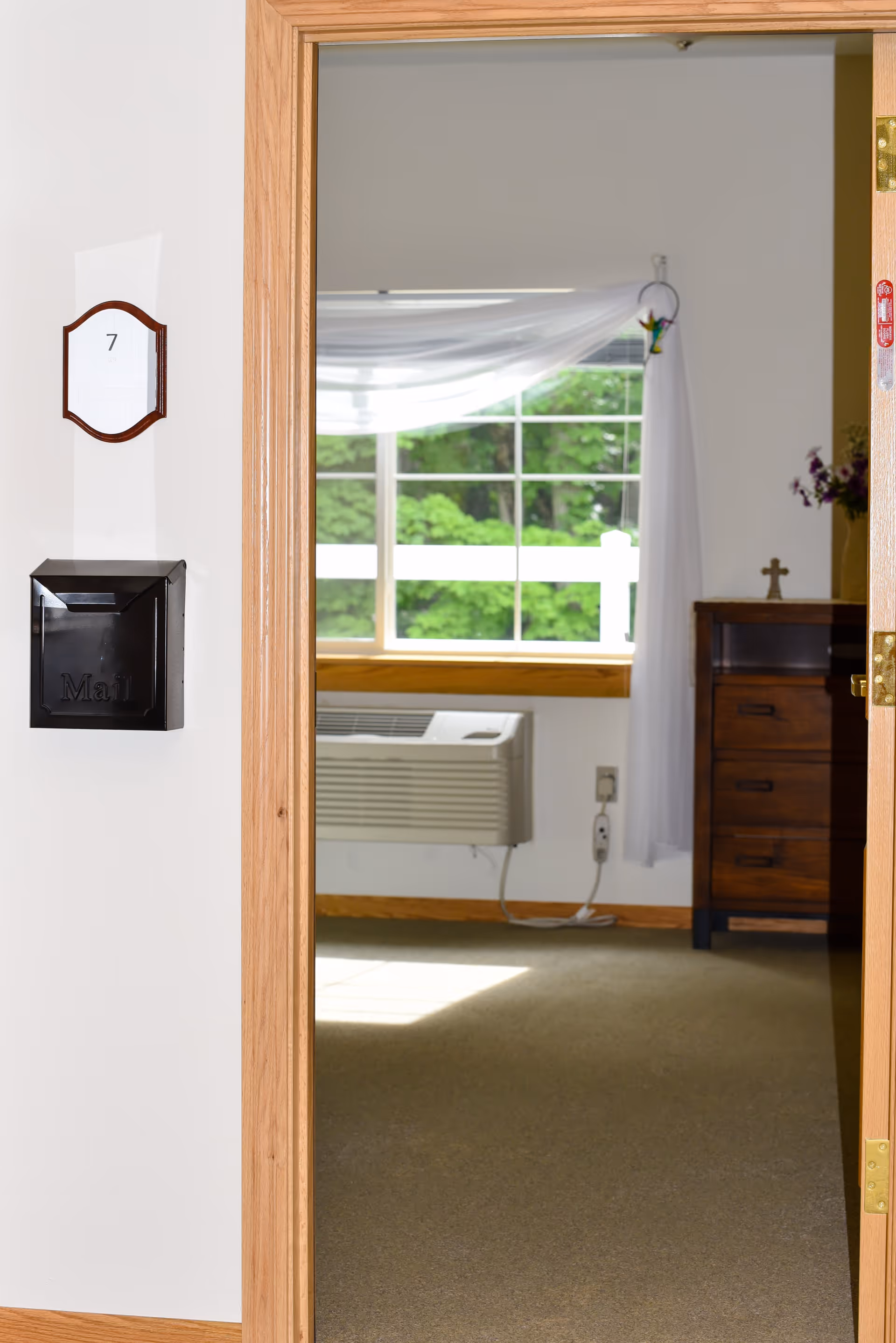 View through an open wooden door into a room with a window showing green trees outside. Inside the room, there is a wooden dresser with a small cross and flowers on top, a white curtain draped over the window, and an air conditioning unit below the window. On the wall outside the door, there is a black mail slot and a room number plaque with the number 7.