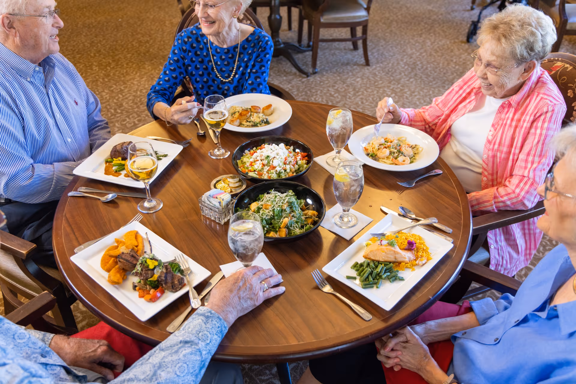 Several elderly residents seated around a round dining table enjoying plated meals and salads in a communal dining room.