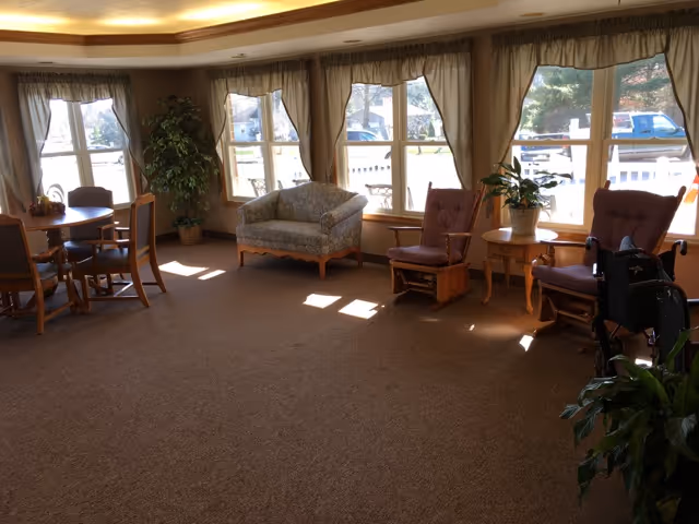 A well-lit common area with large windows covered by beige curtains, featuring a small sofa, two armchairs, a round table with four chairs, several potted plants, and a wheelchair near one of the armchairs.