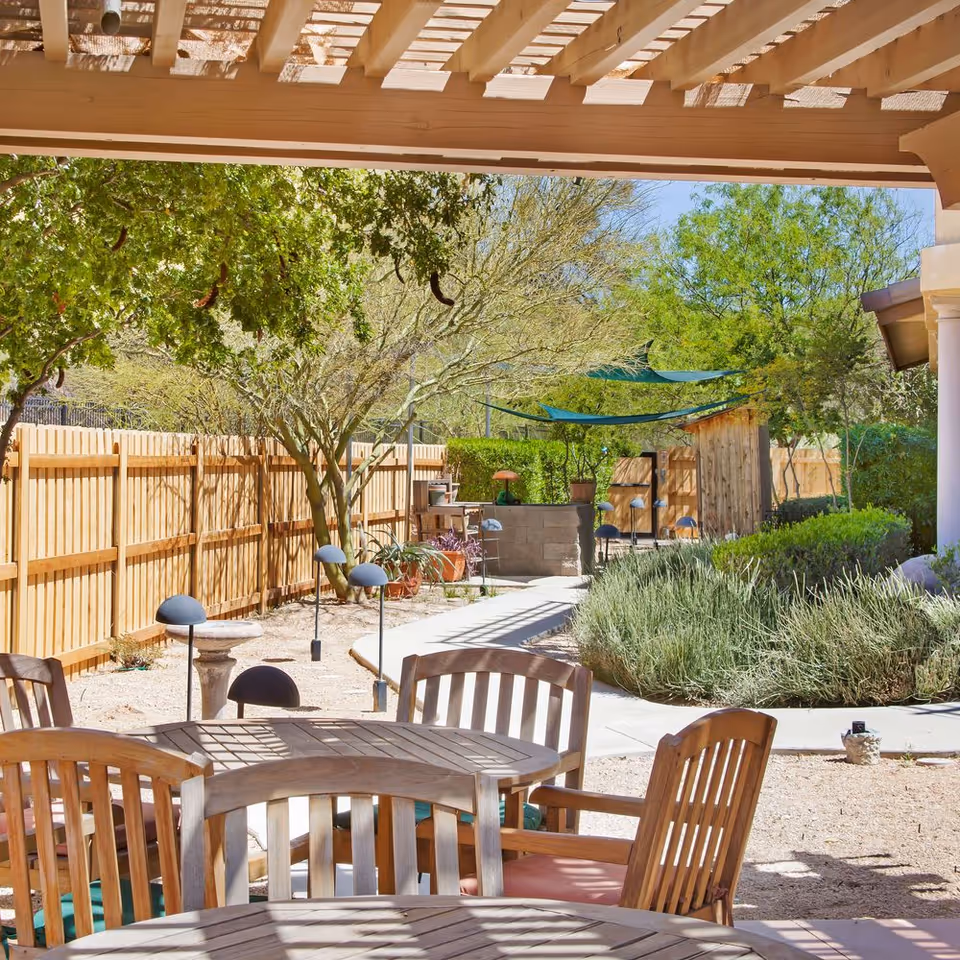 Outdoor patio area with wooden tables and chairs under a pergola, surrounded by trees, shrubs, and a wooden fence. There are shaded seating areas and a paved walkway leading through the garden.