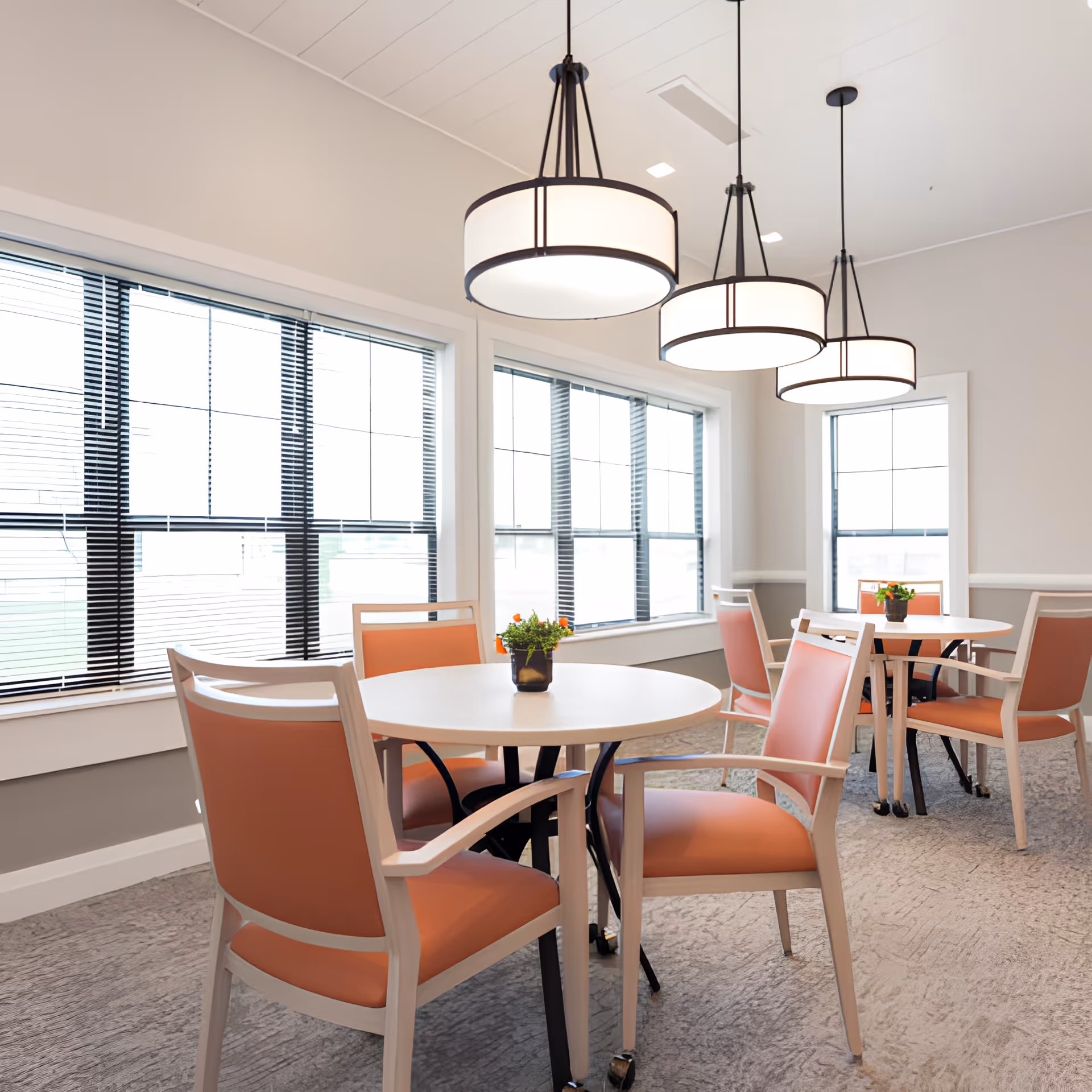 A bright dining area with round white tables and peach-colored cushioned chairs. Three modern pendant lights hang from the ceiling. Large windows with black blinds allow natural light to fill the room. Small potted plants are placed on each table.