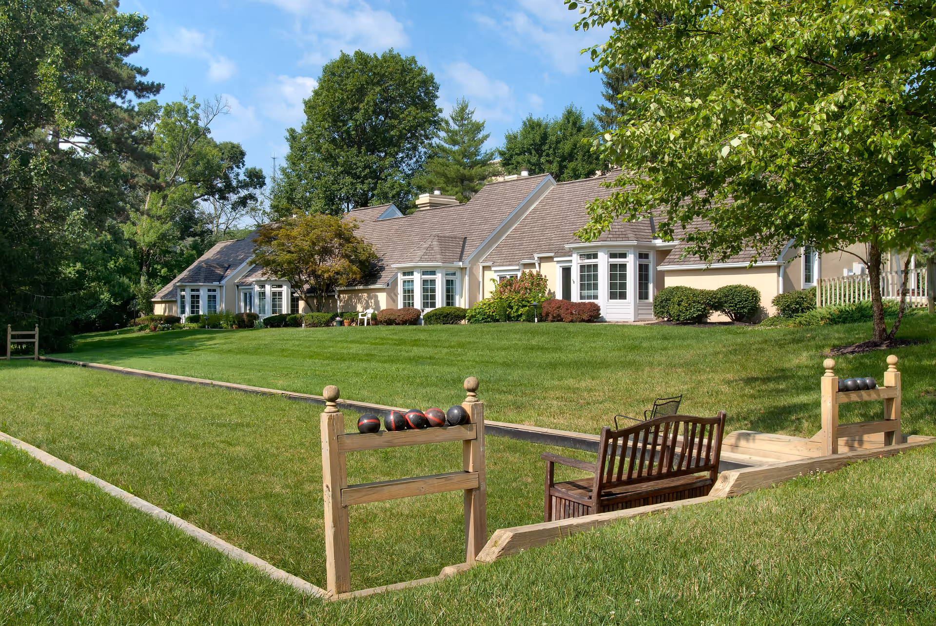 A well-maintained outdoor bocce ball court with wooden benches and racks holding bocce balls, set on a lush green lawn in front of a beige retirement community building surrounded by trees and shrubs under a blue sky.