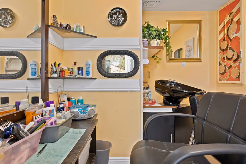 Interior of a hair salon area with a black salon chair, a black hair washing sink, shelves with various hair and beauty products, a mirror, and decorative wall art. The walls are painted yellow and there is a small stereo on the counter.
