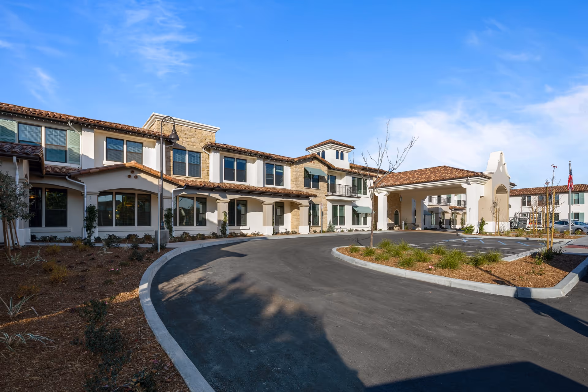 Exterior view of Oakmont of Camarillo senior living facility showing a two-story building with a curved driveway, landscaped areas, and a covered entrance under a clear blue sky.