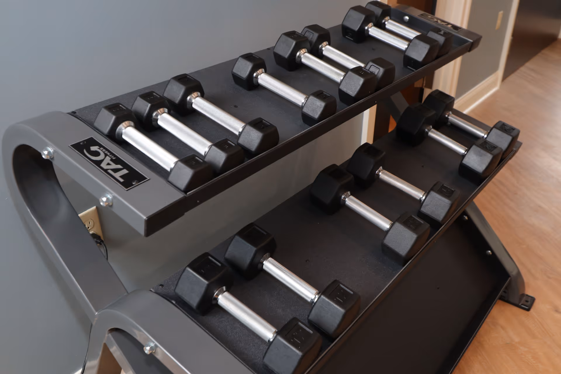 Two-tier rack holding multiple hex dumbbells against a gray wall in a fitness area.