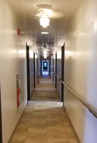 Long indoor hallway with beige tiled floor and white walls, featuring multiple doorways on both sides, ceiling lights, and handrails along the right wall. A fire extinguisher is mounted on the left wall near the foreground.