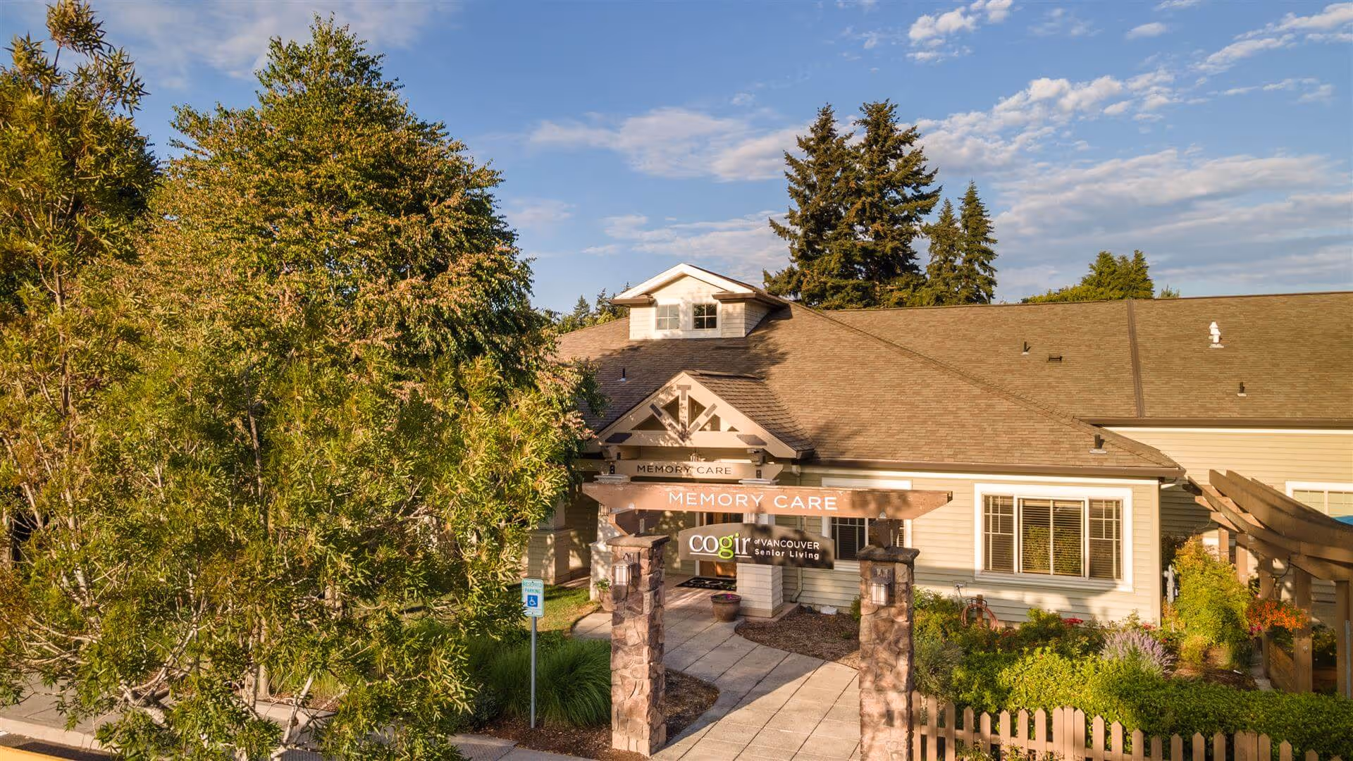 Exterior view of a single-story building with a sign that reads 'Memory Care' and 'Cogir of Vancouver Senior Living' at the entrance. The building is surrounded by trees, bushes, and a wooden fence under a partly cloudy sky.