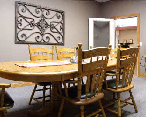 A dining room with a wooden oval table surrounded by six wooden chairs with green cushions. The table has a decorative runner and a small centerpiece. On the wall is a decorative wrought iron wall art. A door with a frosted glass panel is open, showing another room with shelves and framed pictures.