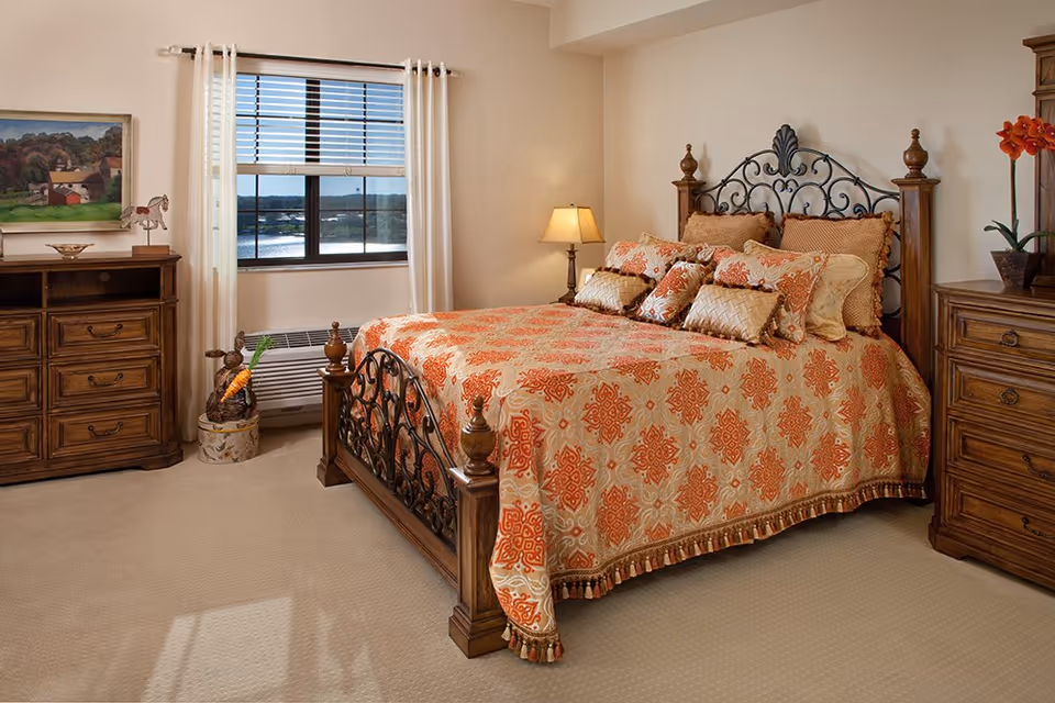 A cozy bedroom with a large bed featuring an ornate metal and wood headboard and footboard. The bed is covered with an orange and beige patterned bedspread and multiple decorative pillows. There are two wooden dressers on either side of the bed, one with a potted plant with orange flowers and the other with a framed painting above it. A window with white curtains and blinds lets in natural light, and a table lamp is on a nightstand next to the bed.