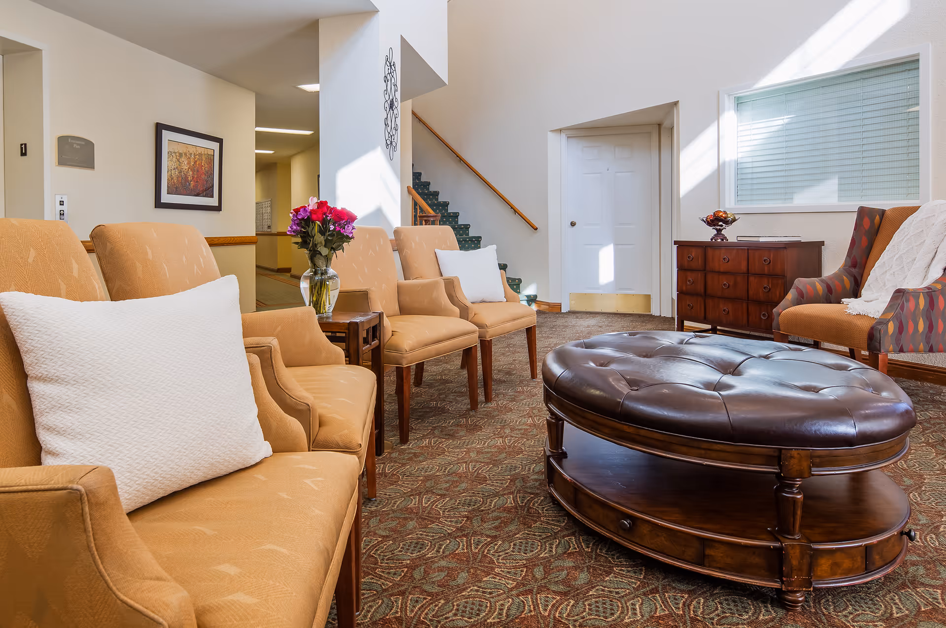A bright and cozy seating area in a senior living facility with several beige armchairs, a large dark brown tufted ottoman in the center, a small wooden side table with a vase of flowers, a wooden dresser with a decorative bowl, and a staircase in the background.