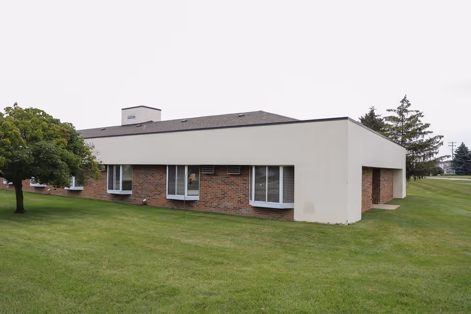 Single-story brick-and-stucco assisted living building on a grassy lawn with windows and a small tree.