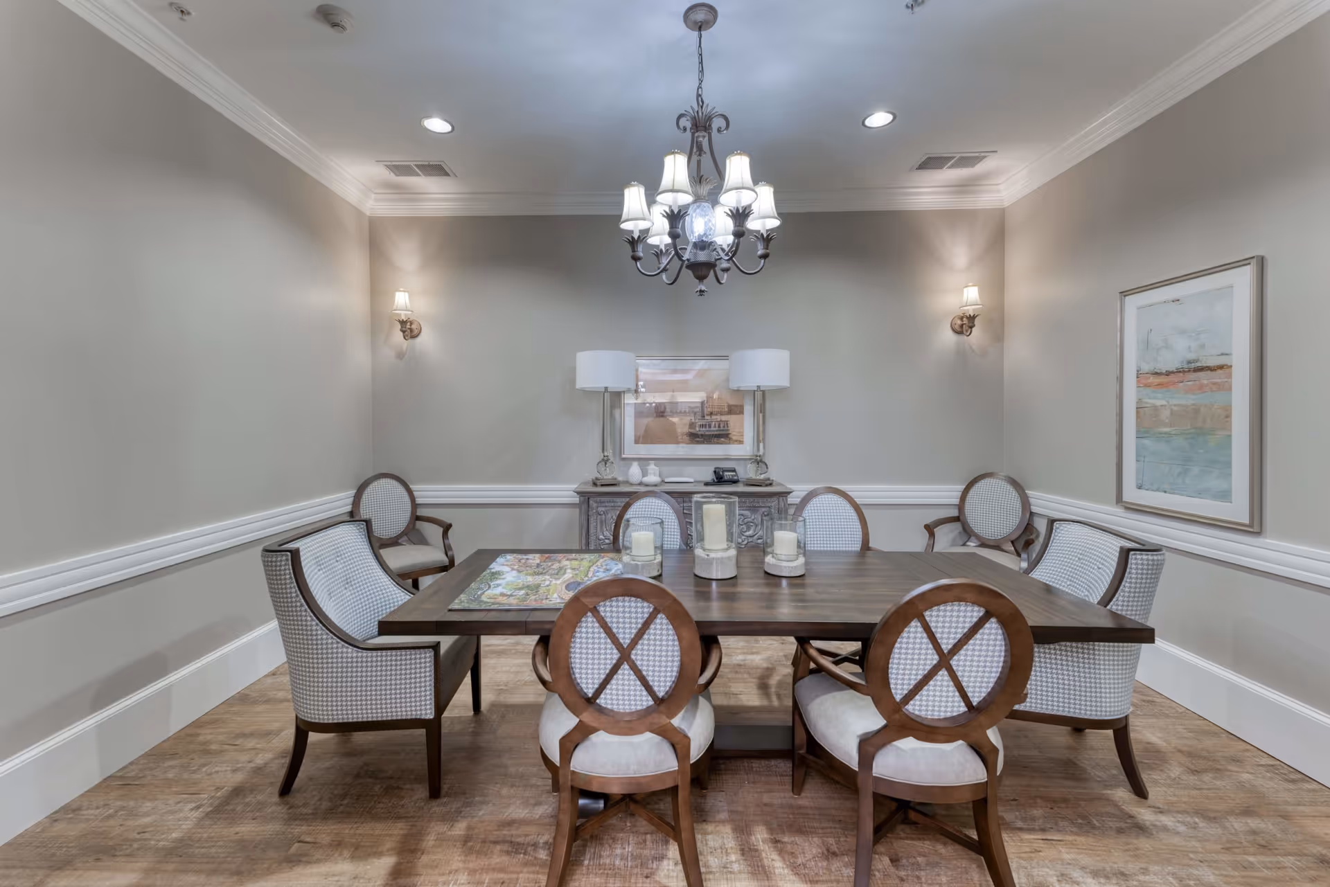 A formal dining room with a rectangular wooden table surrounded by eight upholstered chairs with wooden frames. The room features light gray walls with white trim, a chandelier hanging from the ceiling, two wall sconces, a sideboard with two lamps, decorative items, and a framed picture above it. There is also a framed abstract painting on the right wall and wooden flooring.