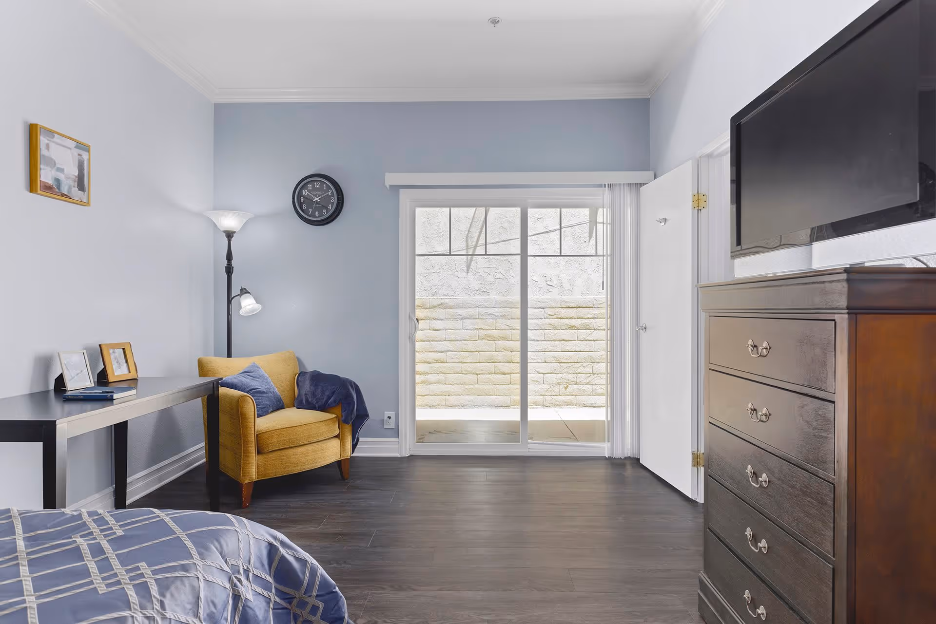 A bedroom with a bed partially visible in the foreground, a yellow armchair with a blue pillow and blanket, a black floor lamp, a black table with picture frames, a wall clock, a sliding glass door leading outside, and a dark wooden dresser with a TV on top.