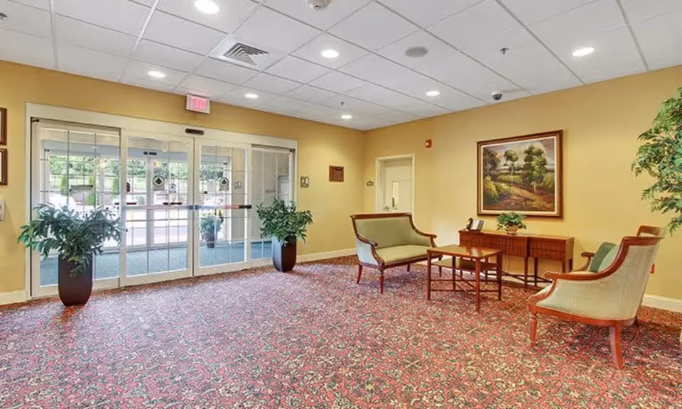 A sitting area inside a senior living facility with a patterned carpet, two potted plants near glass automatic doors, a small wooden table with two chairs and a loveseat, a sideboard with a plant and a framed landscape painting on the wall.