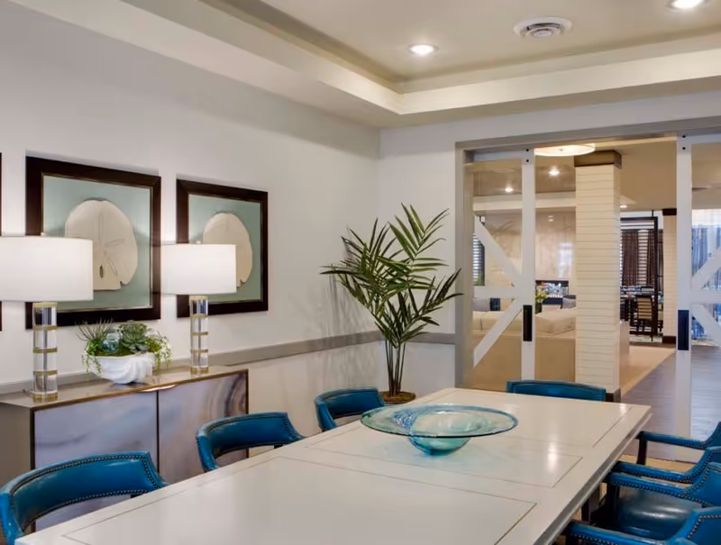Modern dining room with a long white table, teal chairs, a glass centerpiece, wall lamps and framed art opening into a lounge area.
