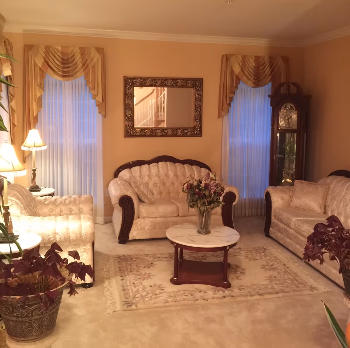 Traditional beige living room with tufted sofas, a marble-top coffee table with flowers, a mirror, lamps, and a grandfather clock between two curtained windows.