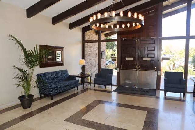 Bright lobby with seating, a circular chandelier, potted plant and double wooden entrance doors with large windows.