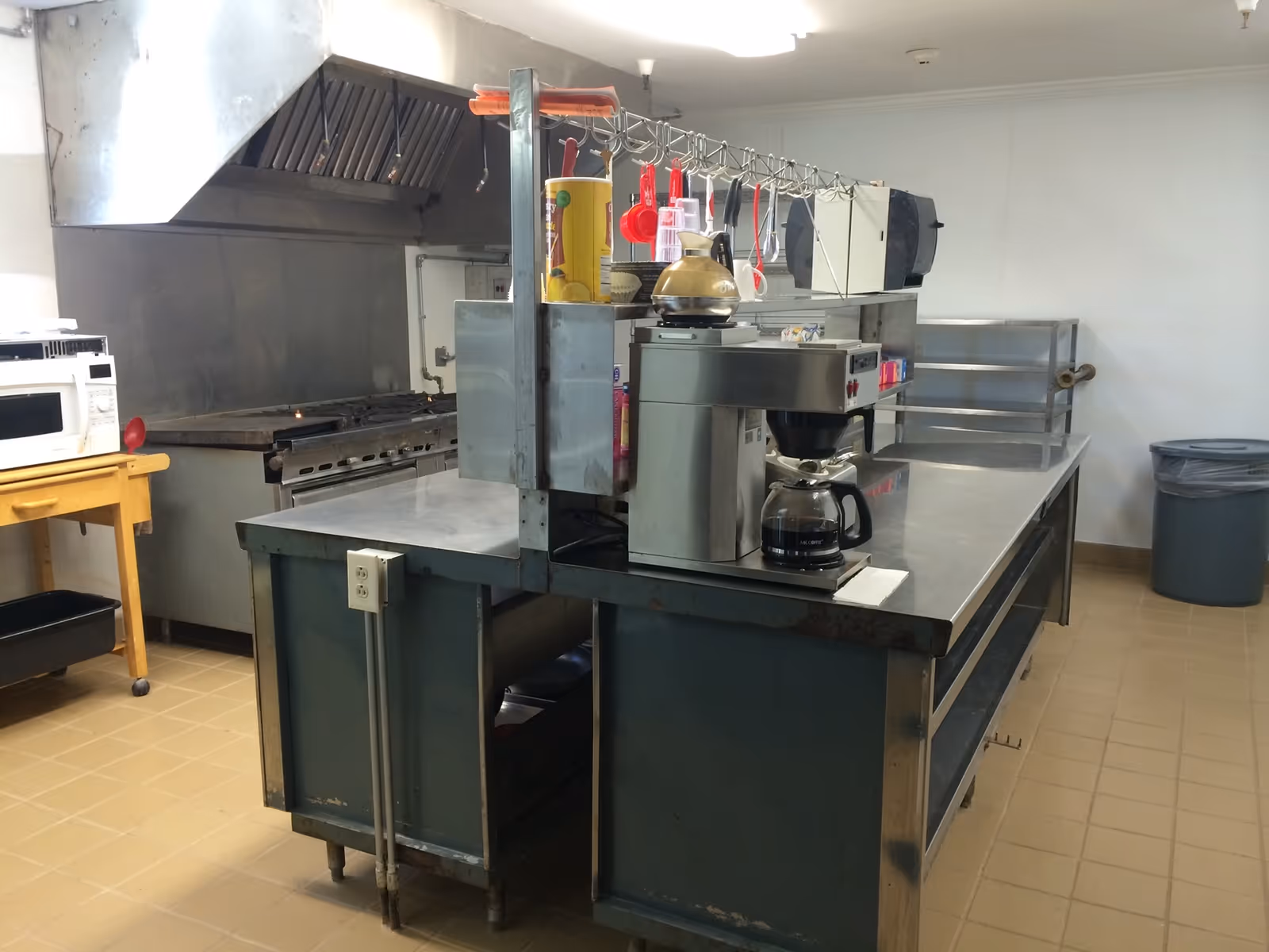 Commercial kitchen area with stainless steel counters, a coffee maker, microwave, stove with exhaust hood, and various kitchen utensils hanging on a rack. There is a trash can in the corner and tiled flooring.