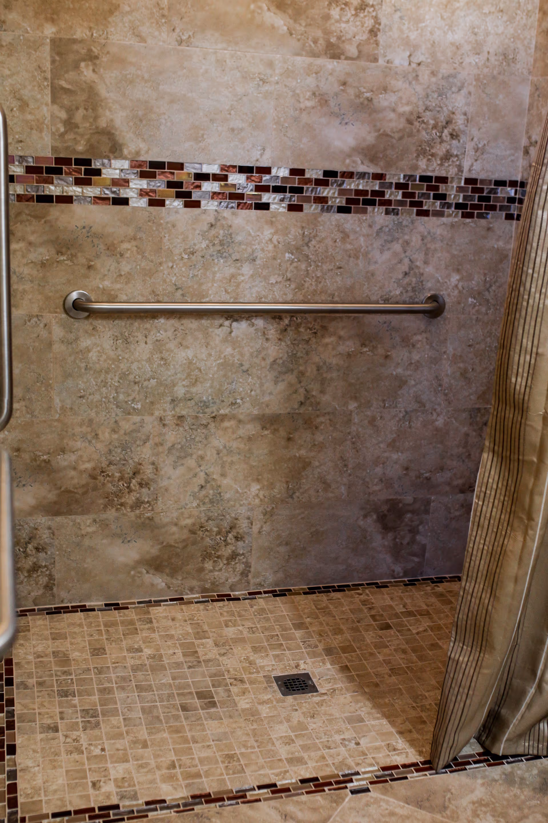 A tiled shower area with beige and brown mosaic tiles on the floor and a decorative horizontal strip of small rectangular tiles in shades of brown, red, and white on the wall. There is a metal grab bar mounted horizontally on the wall and a striped shower curtain partially visible on the right side.
