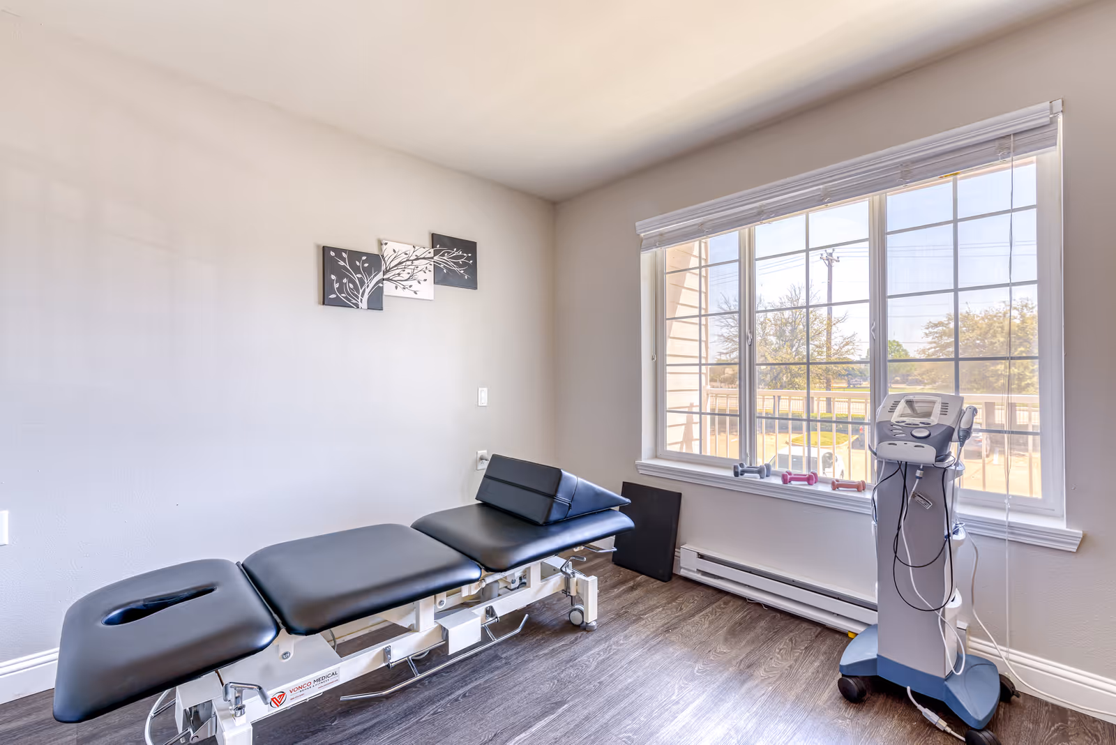 A bright therapy room with an adjustable black treatment table, medical equipment on a cart, and a large window.