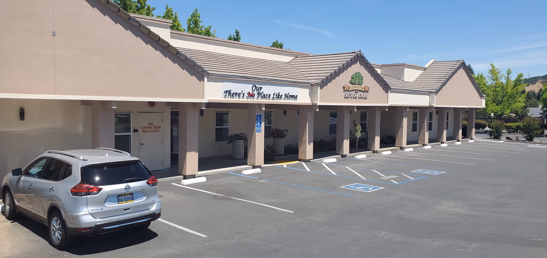 Exterior view of Village Oaks Senior Care building with a beige facade and tiled roof. There is a parking lot in front with several handicap parking spaces and a silver Nissan Rogue parked. The building has a sign that reads 'Village Oaks Senior Care' and another sign that says 'Our There's No Place Like Home'. A person is standing near the entrance under the covered walkway.