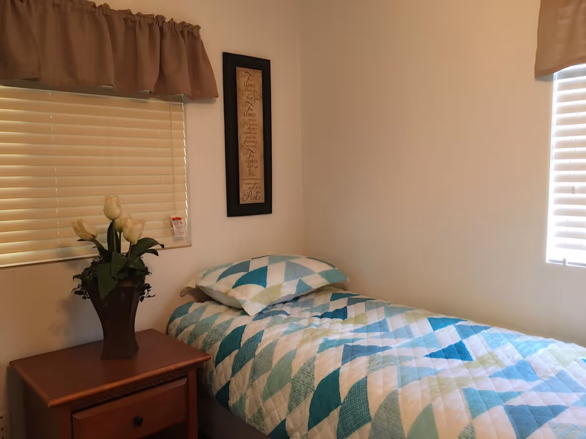 A small bedroom with a single bed covered in a blue, white, and green patterned quilt. Next to the bed is a wooden nightstand with a vase holding white tulips. The room has two windows with beige blinds and matching beige valances. A framed decorative piece with text hangs on the wall above the bed.
