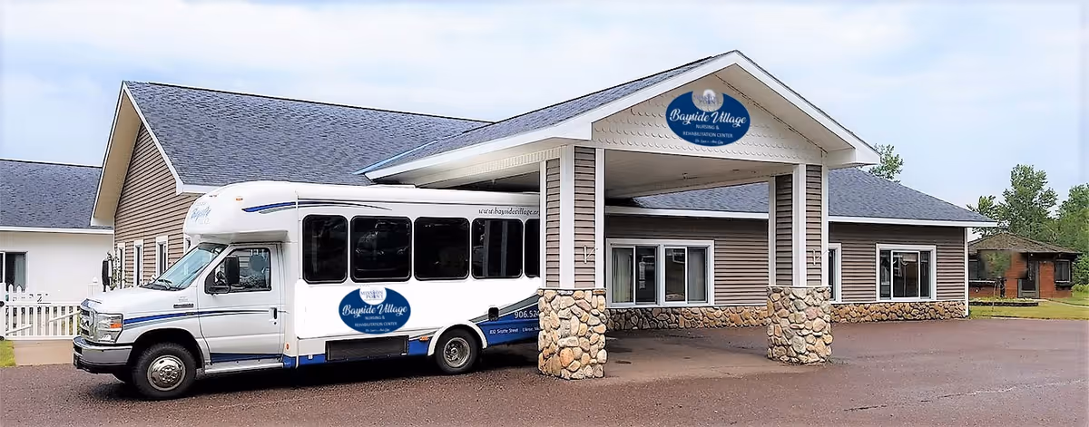 Exterior view of Bayside Village Nursing & Rehabilitation Center showing a single-story building with a covered entrance supported by stone pillars. A white shuttle bus with the facility's logo is parked in front of the entrance.