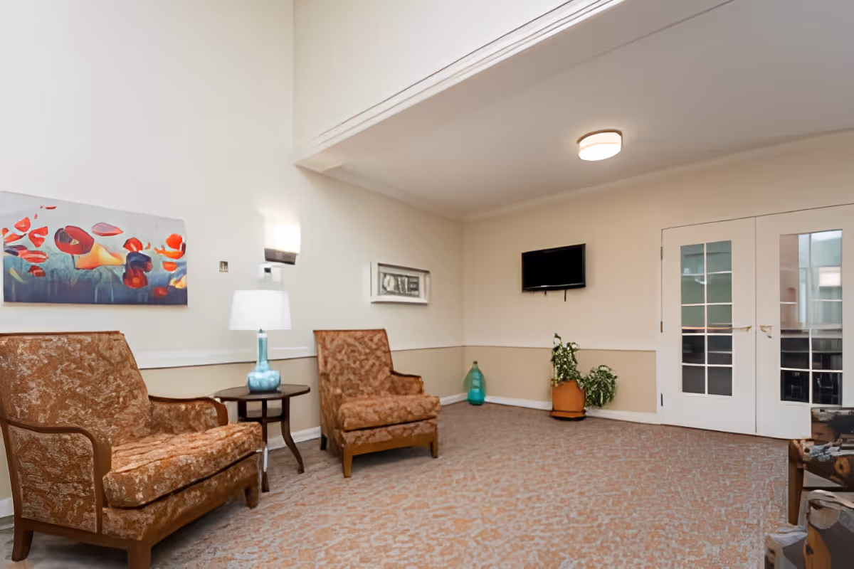 A bright lounge area with two upholstered armchairs, a side table and lamp, wall art, a mounted TV, potted plants and double French doors.