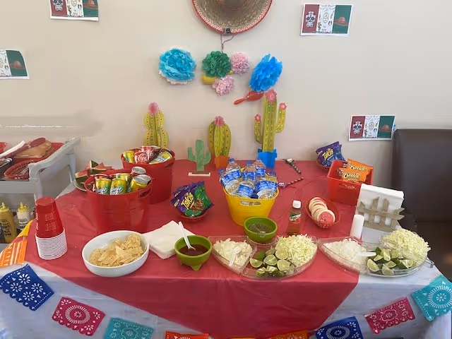 A decorated buffet table with chips, salsas, lime wedges, canned drinks and bottled water set up with cactus and fiesta-themed decorations against a wall.