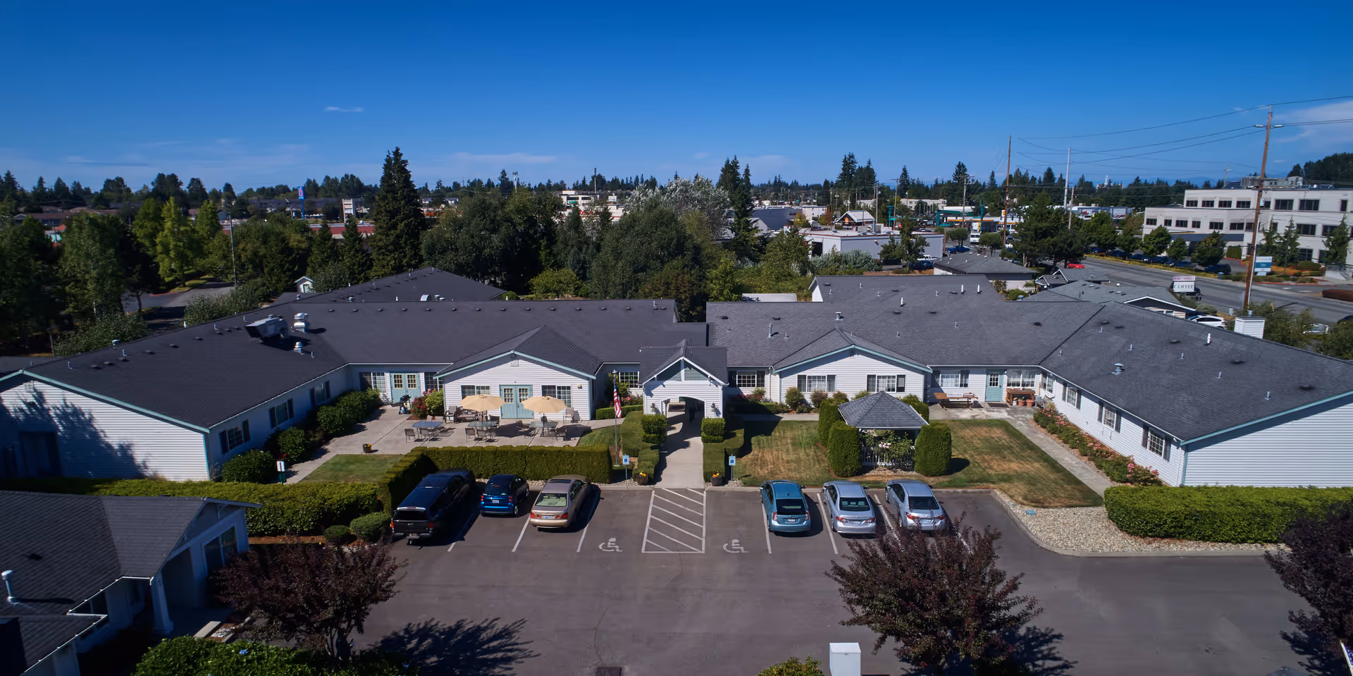 Aerial view of South Pointe Assisted Living facility showing a single-story building with a gray roof and light-colored exterior walls. The building surrounds a central courtyard with green lawns, bushes, and a small gazebo. Several cars are parked in front of the building in designated parking spaces, including accessible parking spots. Trees and other buildings are visible in the background under a clear blue sky.