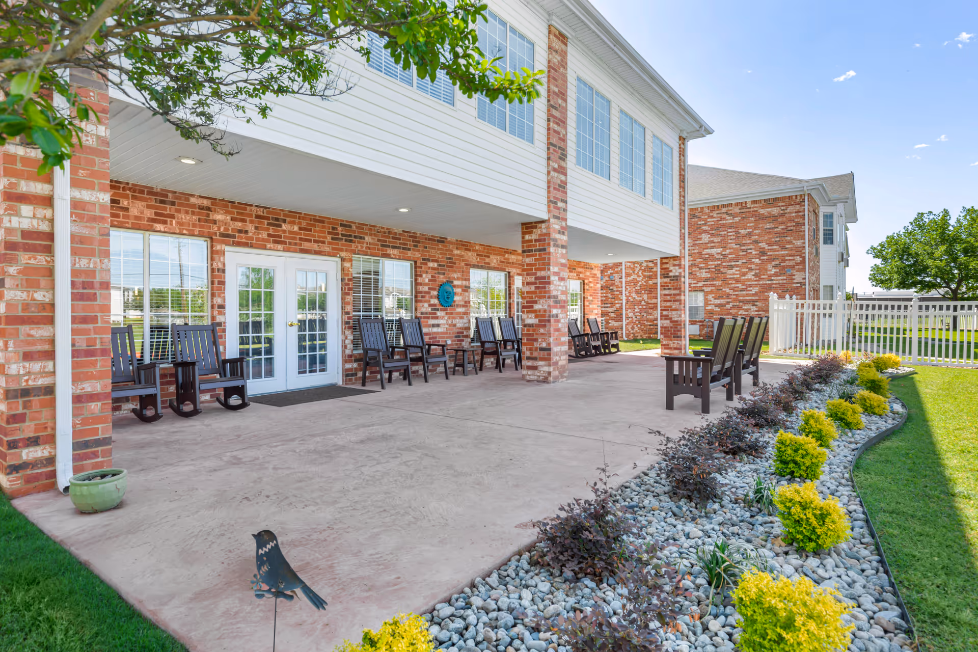 Outdoor patio area of a senior living facility with brick walls and white siding. The patio has several dark wooden rocking chairs and regular chairs arranged along the wall and around the open space. There is a landscaped garden bed with small yellow bushes and decorative rocks bordering the patio. A white fence and green lawn are visible in the background under a clear blue sky.
