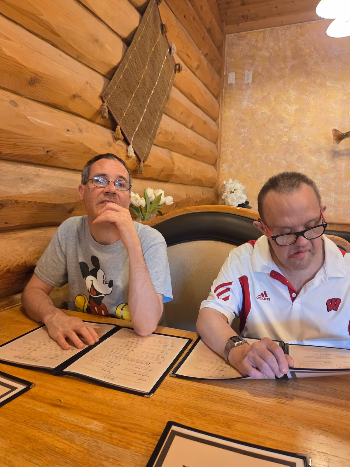 Two men sitting at a wooden table in a cozy room with log cabin walls and a decorative wall hanging. Both men are looking at menus placed on the table in front of them. One man is wearing a gray Mickey Mouse t-shirt and glasses, while the other is wearing a white and red Adidas polo shirt and glasses. There are white flowers in the background on a shelf.