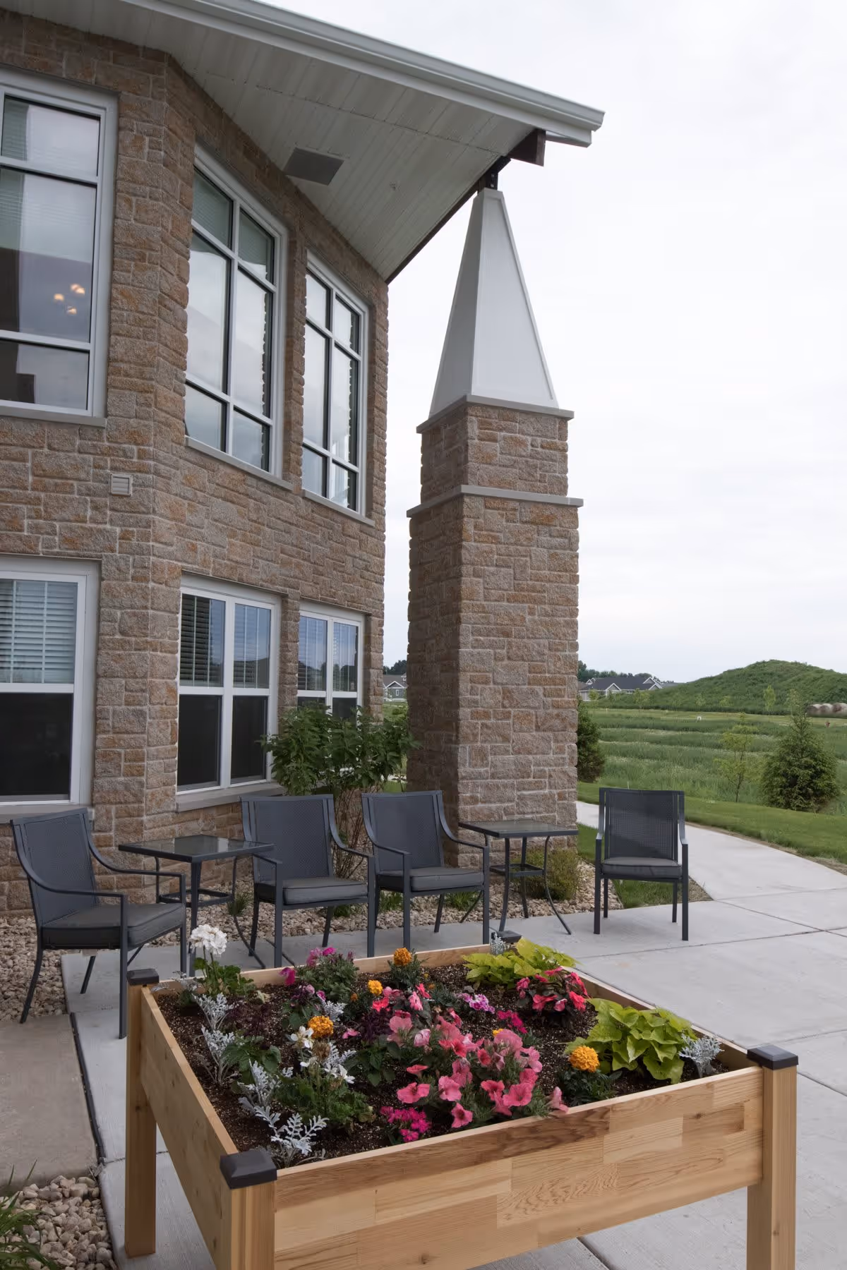Outdoor patio area at a senior living facility with a raised wooden garden bed filled with colorful flowers in the foreground. Behind the garden bed are four black metal chairs and two small tables on a concrete patio next to a stone building with large windows. Green fields and a cloudy sky are visible in the background.