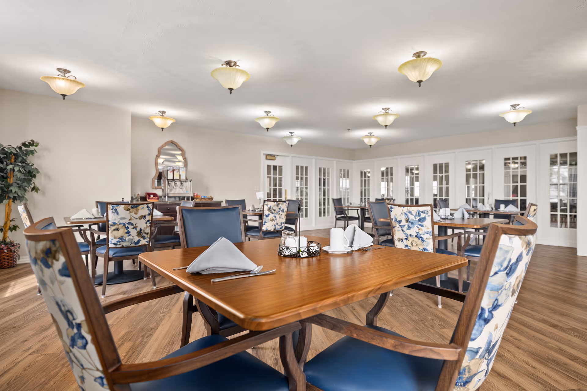 A spacious dining room with wooden tables and chairs featuring floral upholstery. The tables are set with folded napkins, cups, and silverware. The room has wooden flooring, multiple ceiling lights, and white French doors along the back wall. There is a decorative mirror and a beverage station against one wall.