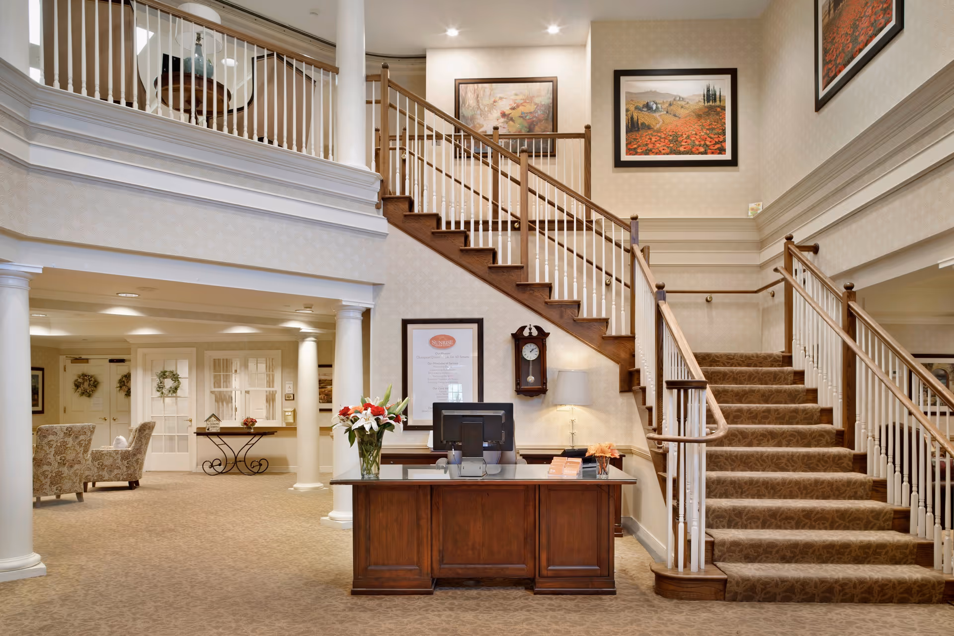 Interior view of a senior living facility lobby featuring a wooden reception desk with a computer and a vase of flowers. Behind the desk is a staircase with wooden handrails and white balusters leading to an upper floor. The walls are decorated with framed paintings and a wall clock. The area has beige patterned carpet and wallpaper, with white columns and a seating area with floral upholstered chairs visible in the background.