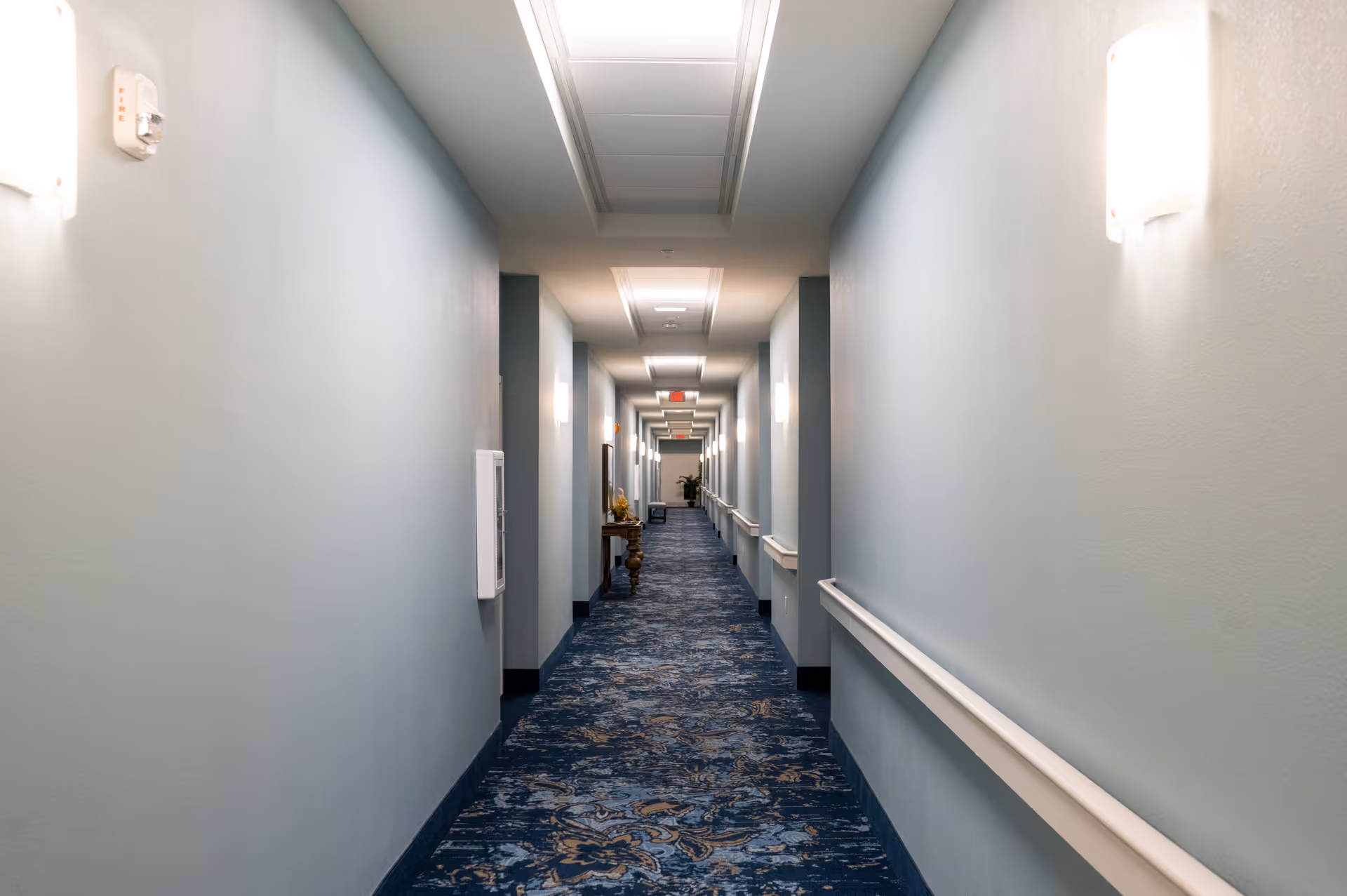 A long, well-lit hallway with light blue walls and a patterned blue carpet. The hallway has white handrails on both sides and several wall-mounted lights. There is a small table with decorative items along the left side and an exit sign visible at the far end.
