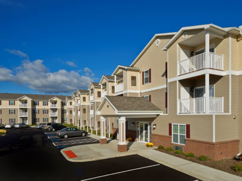 Three-story beige apartment-style senior living building with balconies, a covered main entrance, and a parking lot under a blue sky.