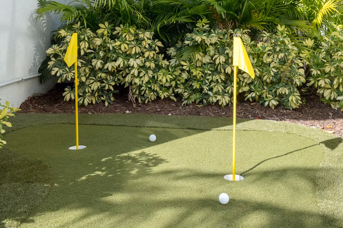 A small outdoor putting green with two yellow flags and two golf balls on artificial grass, surrounded by green leafy plants and a white wall.