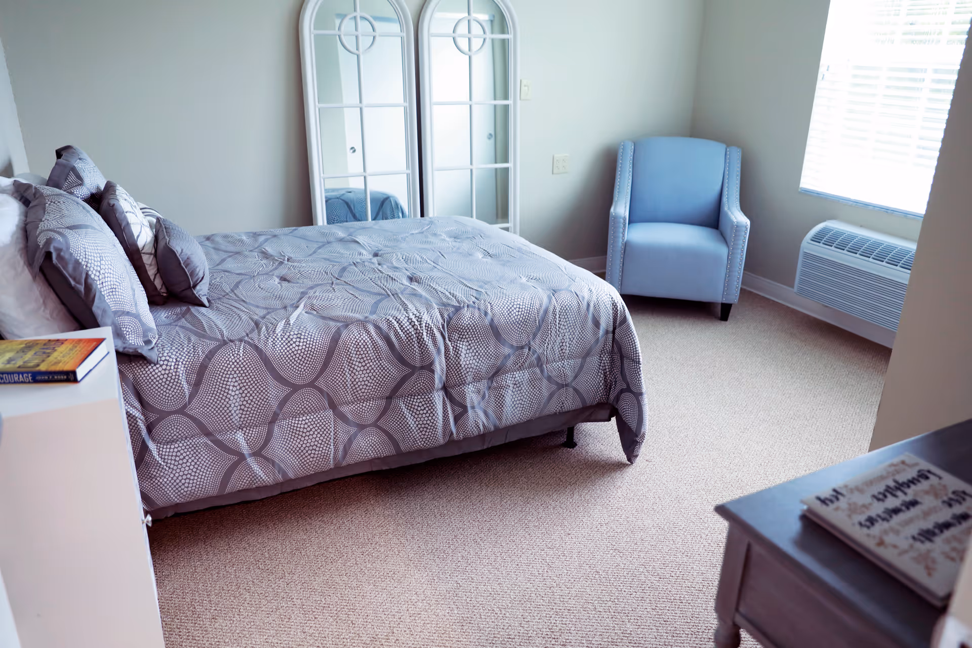 A bedroom with a bed covered in a patterned gray and white comforter, several pillows, a blue upholstered armchair near a window with blinds, a white dresser with a book on top, and a large white-framed mirror leaning against the wall.
