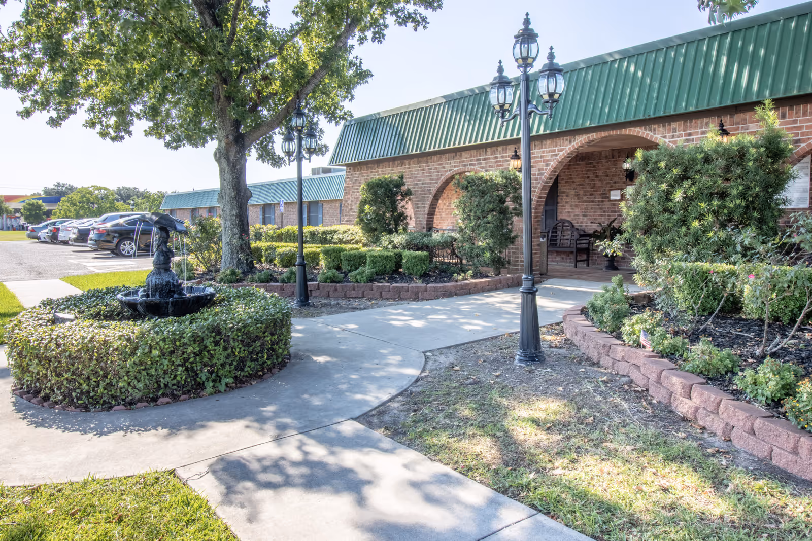 Outdoor view of San Jacinto Manor showing a brick building with green metal roofing, a paved walkway, a black fountain surrounded by greenery, lamp posts, and parked cars in the background under a clear sky.