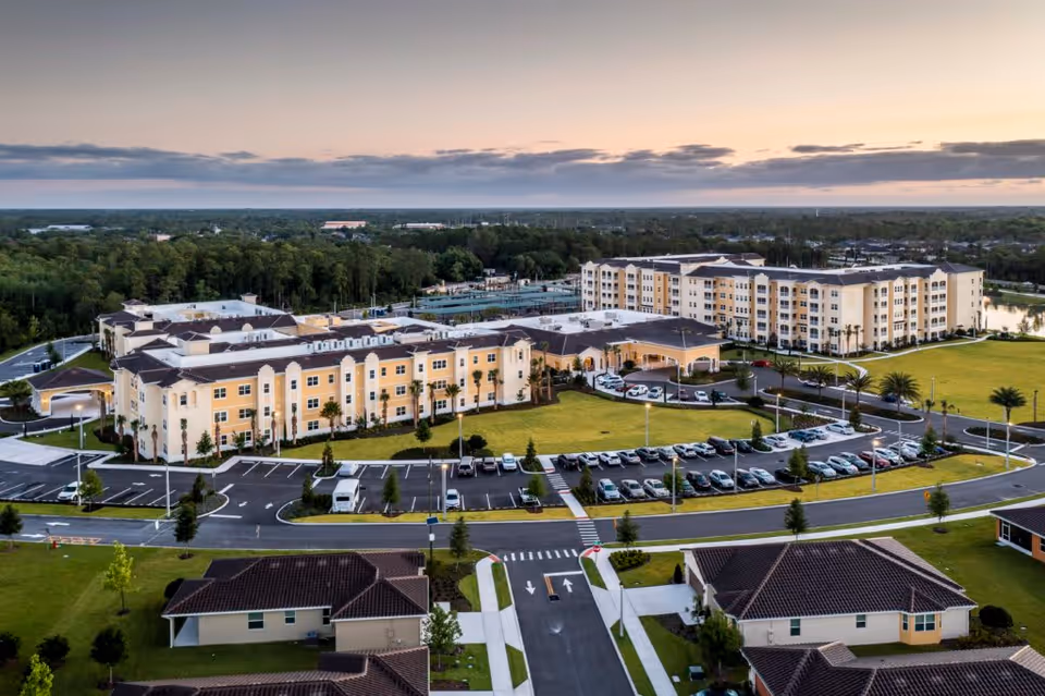 Aerial view of Legacy Pointe at UCF, showing a large senior living facility with multiple connected buildings, parking lots filled with cars, landscaped green lawns, and surrounding residential houses under a partly cloudy sky at sunset.