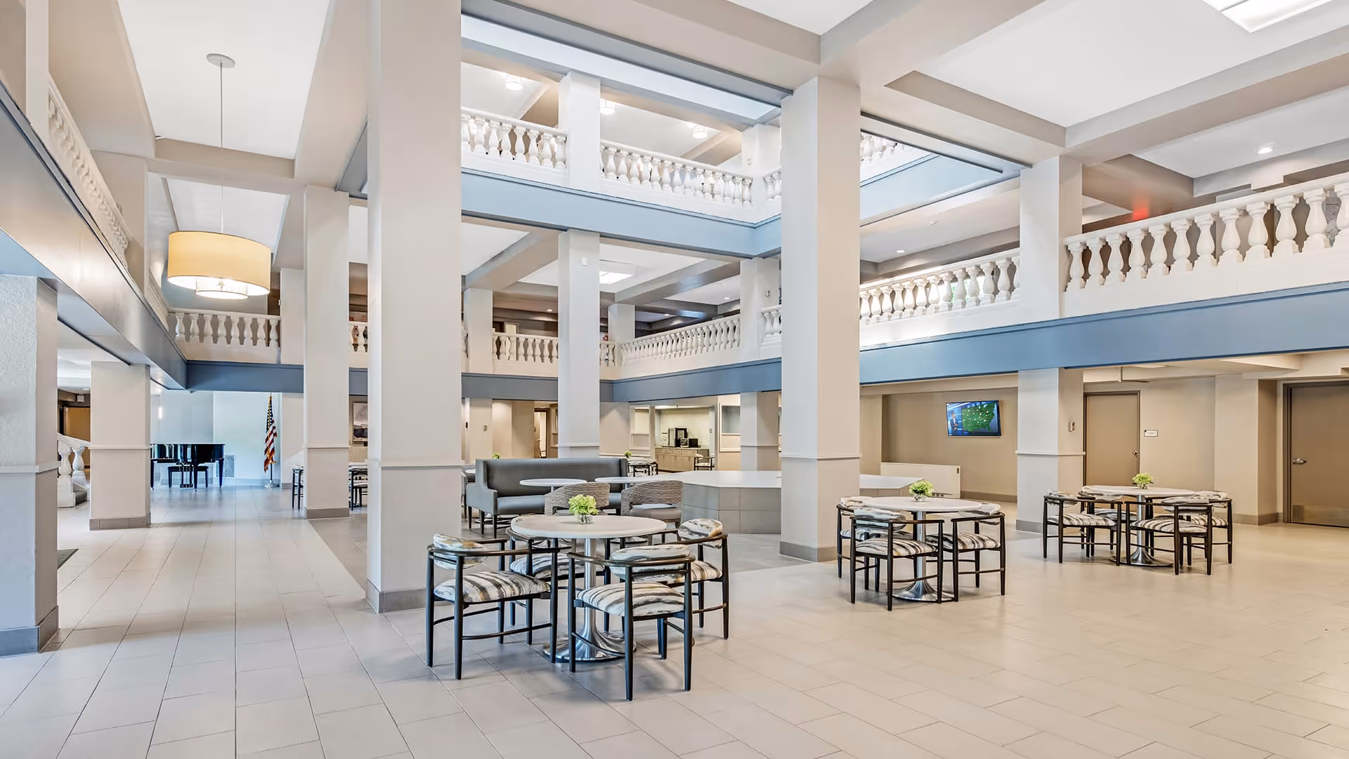 Spacious and bright common area in a senior living facility with multiple round tables and chairs arranged for seating. The room features high ceilings with large pillars, a second-floor balcony with decorative railings, and a piano in the background. The area is well-lit with natural and artificial lighting, and there is a television mounted on the wall.