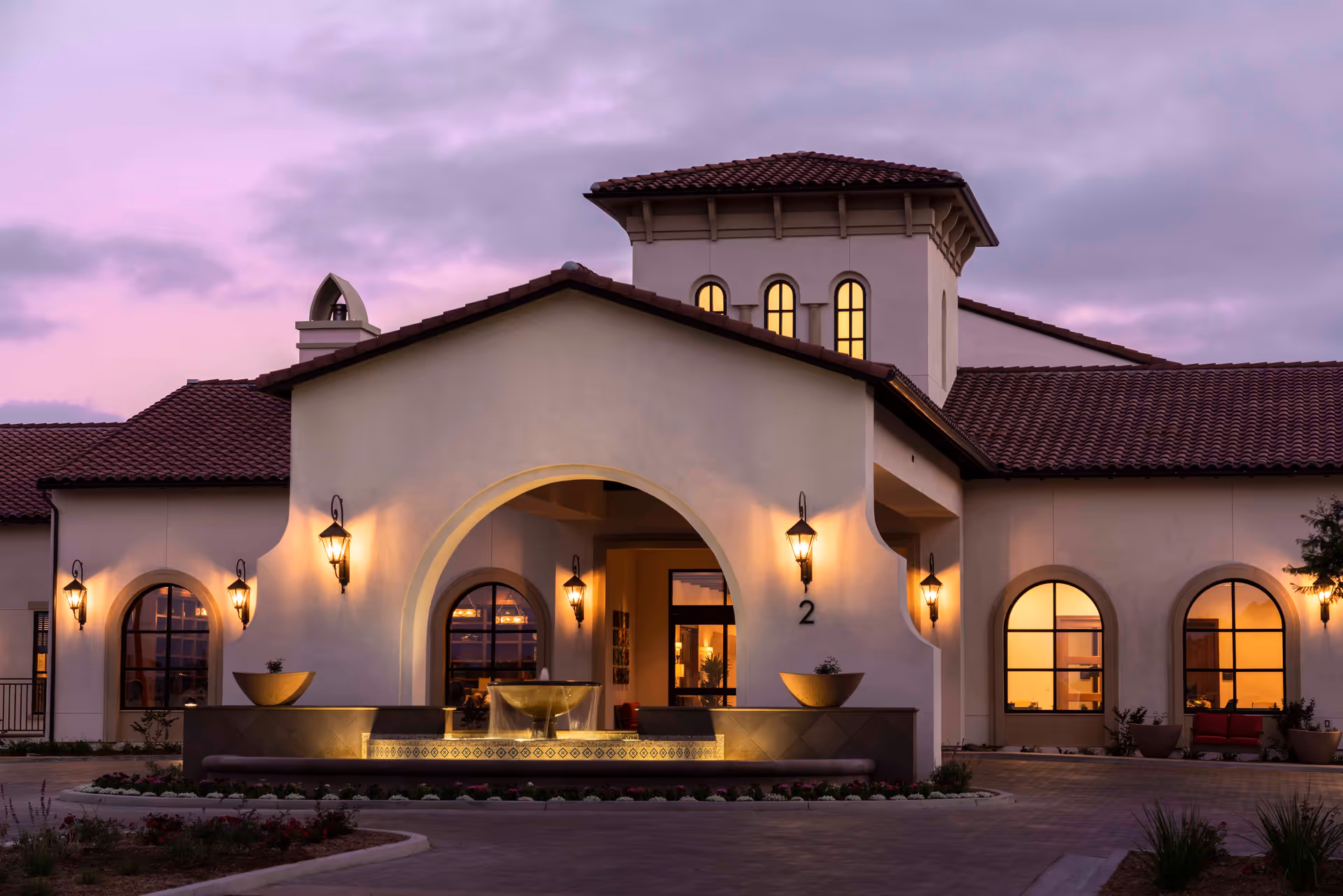 Exterior view of a senior living facility building at dusk with warm lights glowing through arched windows and wall-mounted lanterns. The building features a Mediterranean architectural style with a tiled roof, an arched entrance, and a decorative water fountain in front.