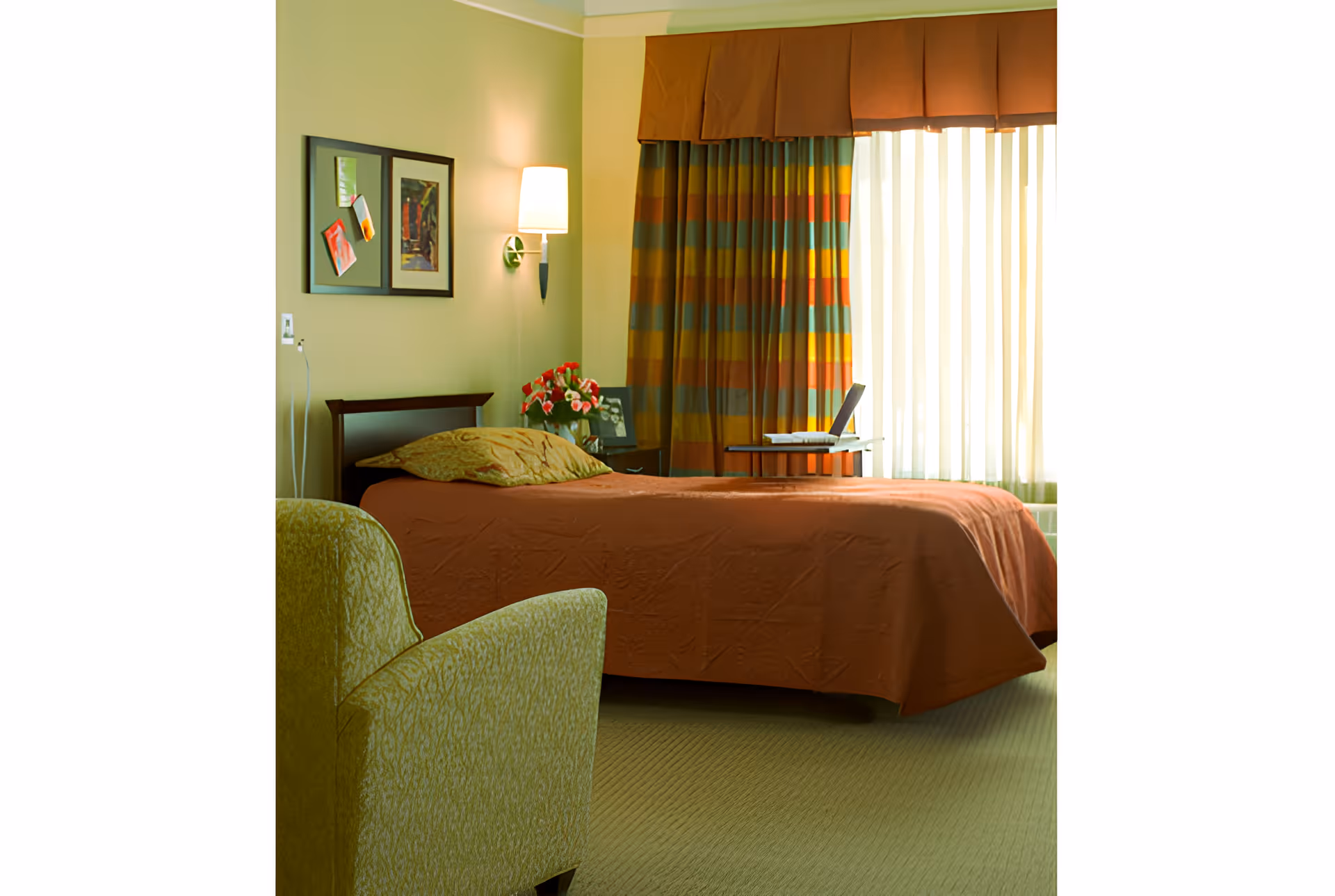 A cozy bedroom with a single bed covered in a rust-colored bedspread and a matching pillow. Next to the bed is a small table with a laptop and a vase of red flowers. A green upholstered armchair is positioned in the foreground. The room has light green walls, two framed pictures on the wall, a wall-mounted lamp, and colorful striped curtains covering a large window.