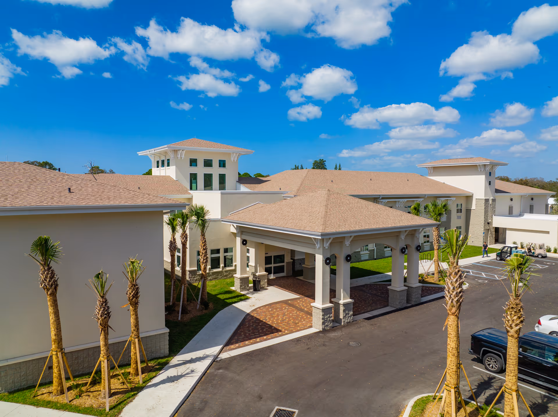 Exterior view of Pines of Sarasota Health Care, LLC building with a covered entrance, palm trees, parked vehicles, and a bright blue sky with scattered clouds.