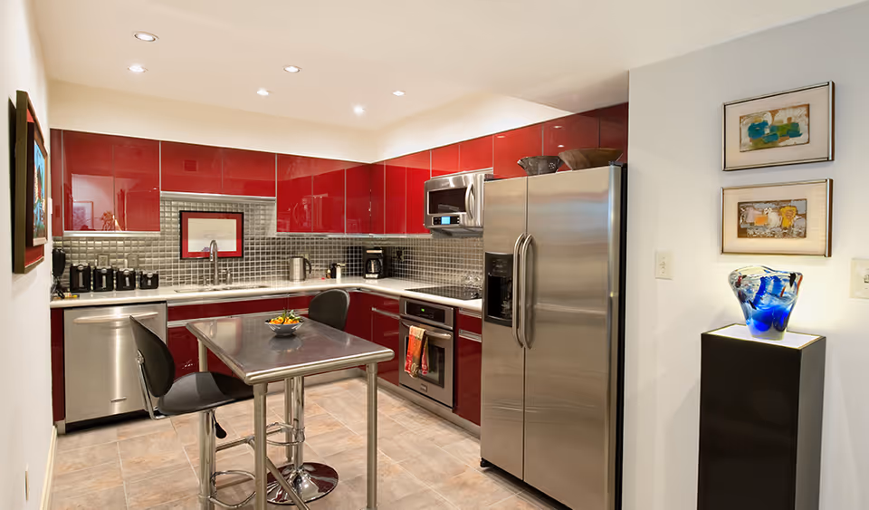Modern kitchen with glossy red cabinets, stainless steel appliances including a refrigerator, oven, microwave, and dishwasher. A small metal table with two black chairs is in the center. The backsplash is made of small gray tiles, and the floor is tiled. On the right side, there is a pedestal with a blue decorative glass piece and two framed artworks on the wall above it.